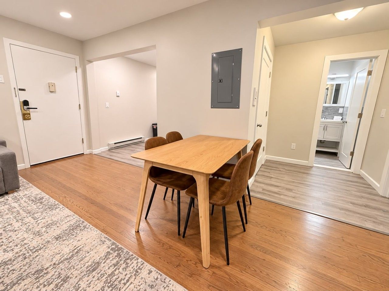 A dining area with a wooden table and chairs, a patterned rug, and a kitchen visible through an open door.
