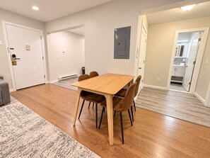 A dining area with a wooden table and chairs, a patterned rug, and a kitchen visible through an open door.