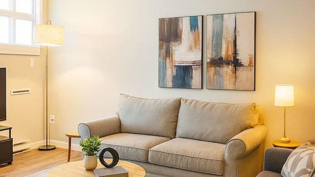 Una sala de estar moderna con un sofá, un sillón y una mesa de centro. Hay dos cuadros abstractos en la pared y una lámpara de pie.