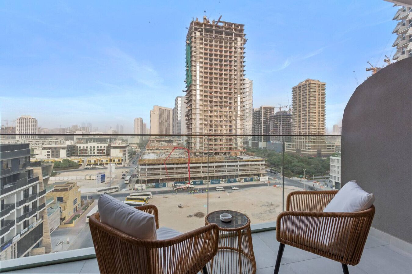 A balcony with wicker chairs and a small table, overlooking a cityscape with high-rise buildings and construction sites.
