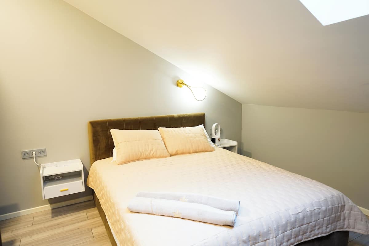 A neatly made bed with a headboard, a bedside table with a lamp, and a power outlet on the wall.