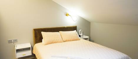 A neatly made bed with a headboard, a bedside table with a lamp, and a power outlet on the wall.