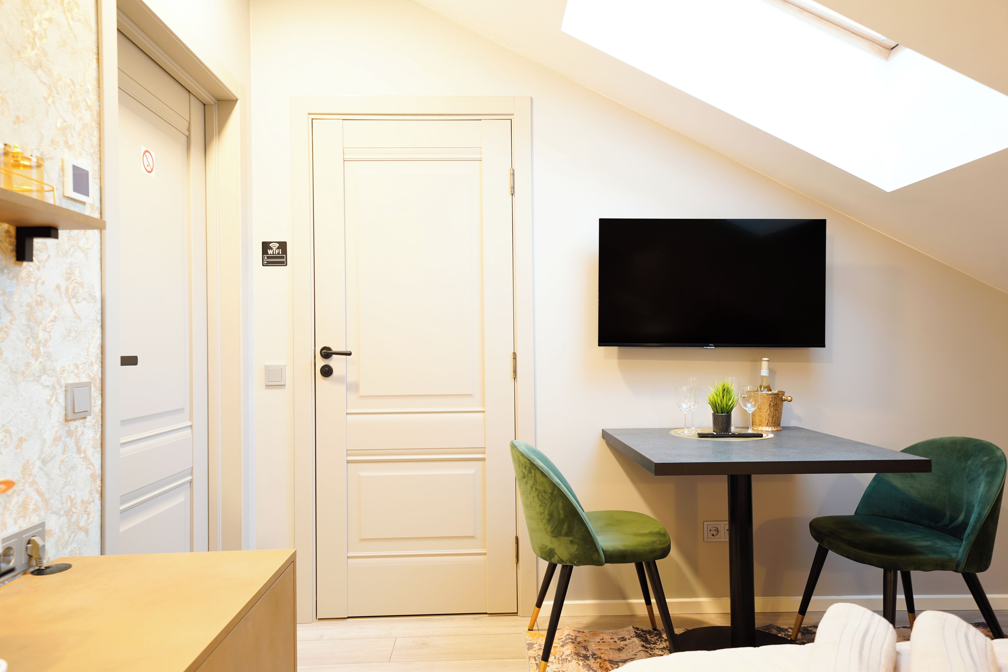 Ein modernes Zimmer mit einem an der Wand montierten Flachbildfernseher, einer weißen Tür mit Rauchverbotsschild und zwei grünen Sesseln um einen kleinen Tisch mit einer Pflanze und einer Flasche.