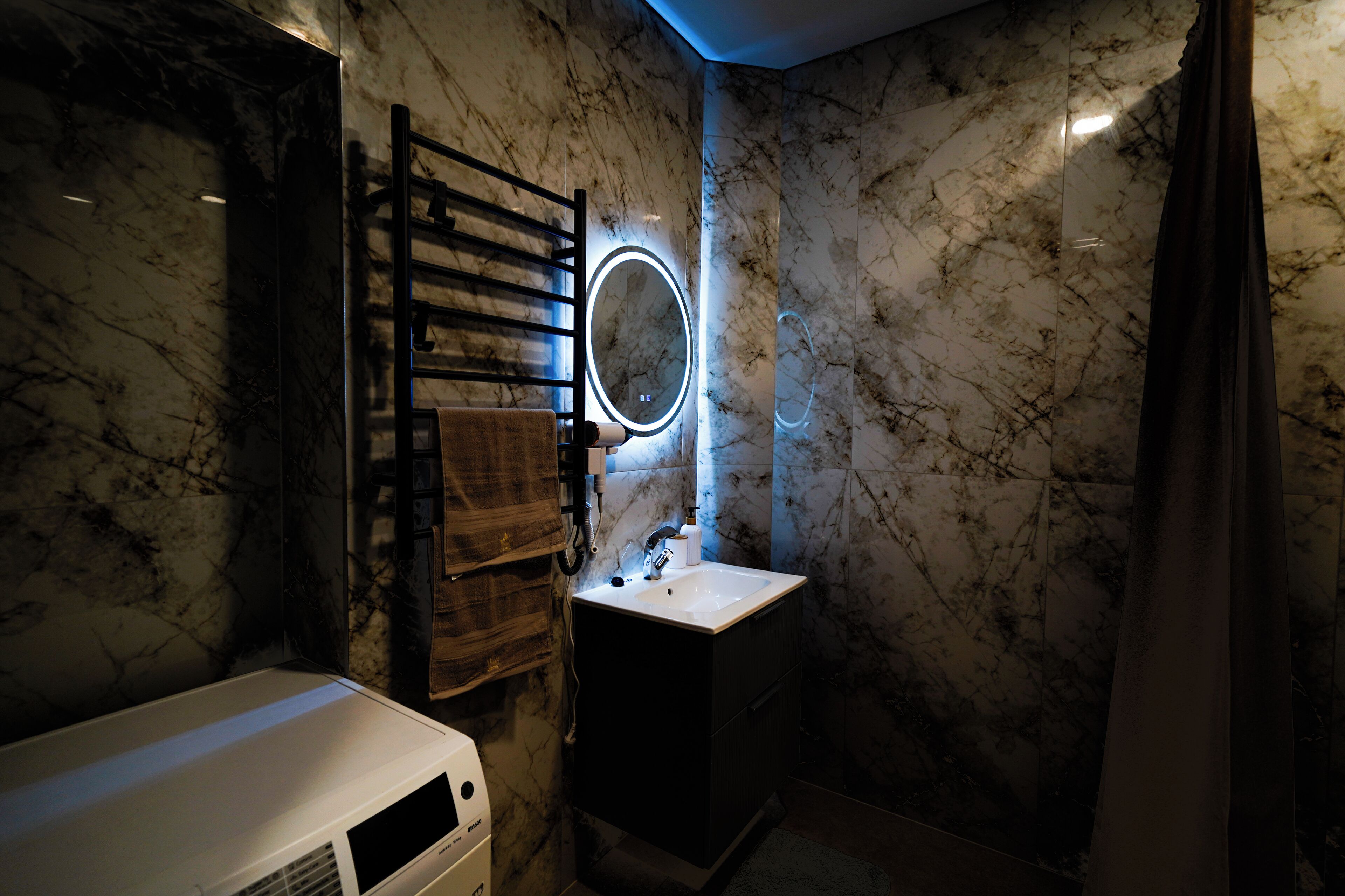 A bathroom with a marble wall, a wooden towel rack, a sink with a mirror, and a towel dispenser.