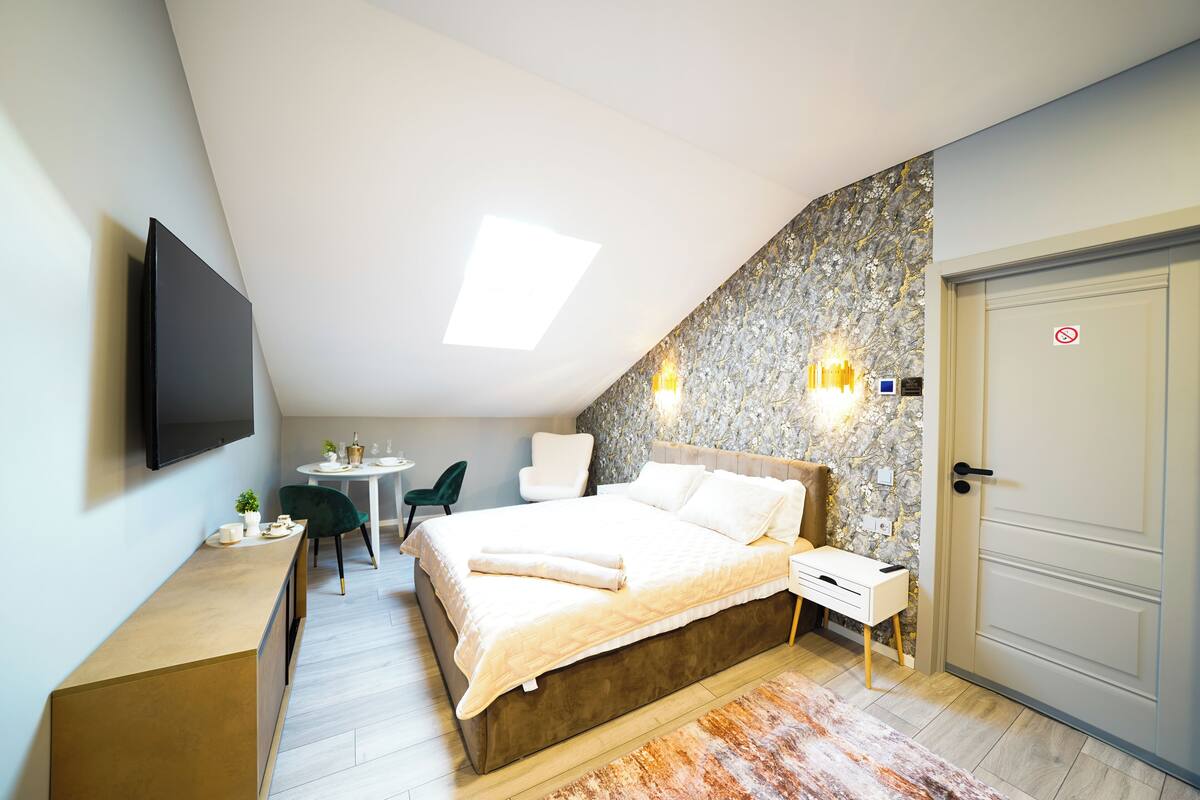 A modern bedroom with a sloped ceiling, a bed with a beige duvet, a desk with a chair, a small table with a plant, and a wall-mounted TV.