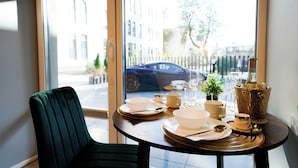 A dining table set with plates, bowls, and a gold bucket, positioned near a window with a view of a street and parked car. - Svaja ApartHotel & Spa (Šiauliai)