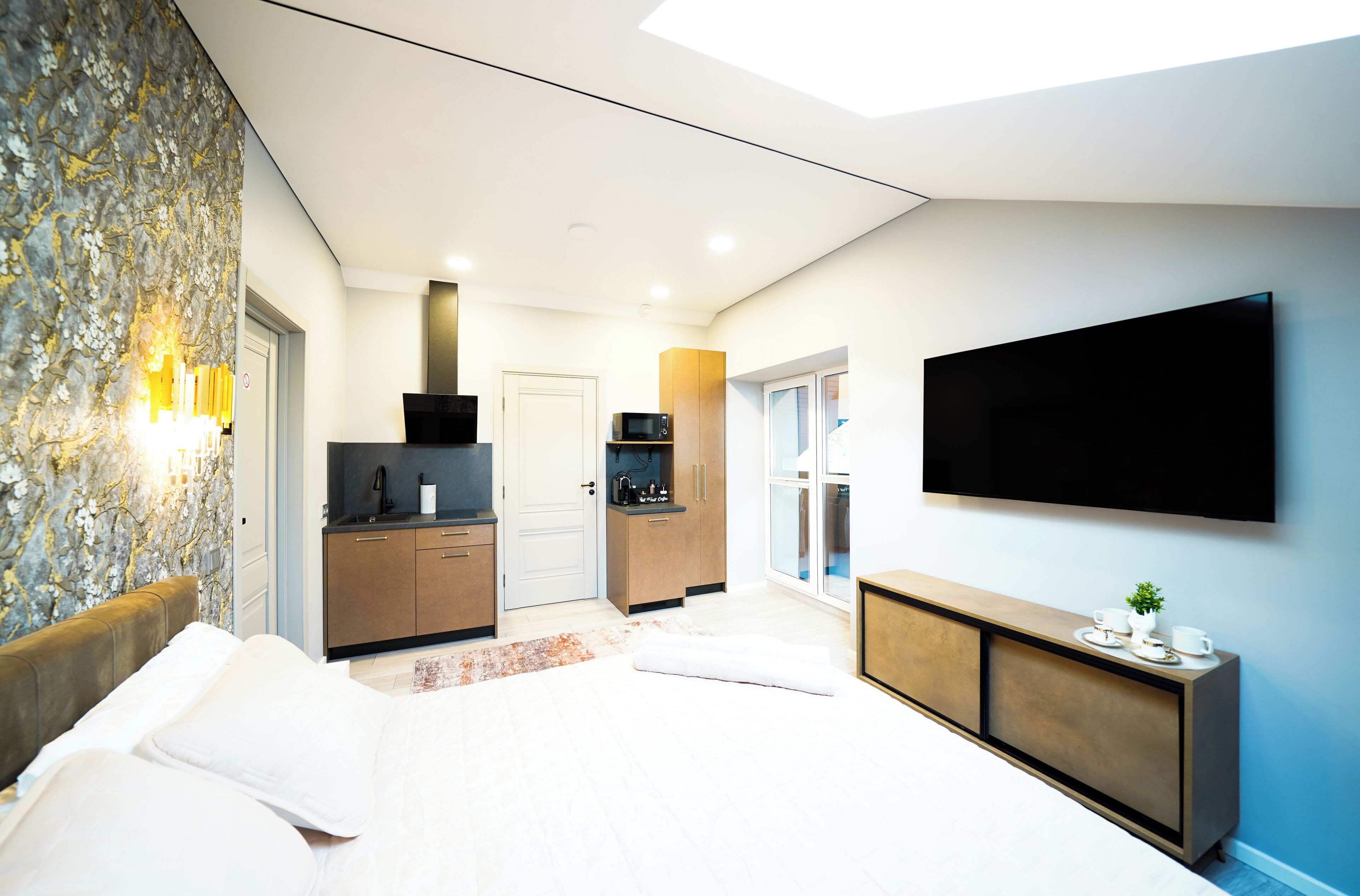 A modern hotel room with a large bed, a flat-screen TV mounted on the wall, a kitchenette, and a stone-textured accent wall.