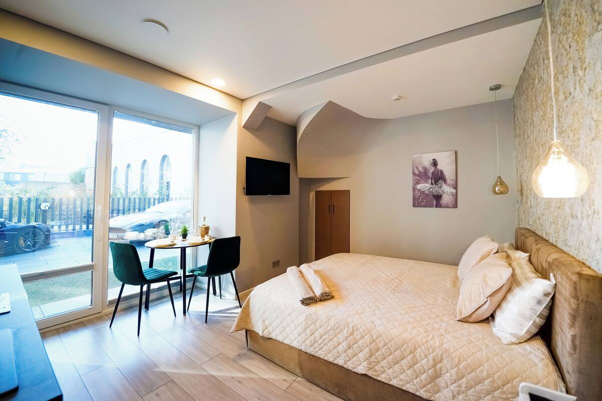 A modern bedroom with a large bed, a dining area with chairs, a table, and a view of the outside.