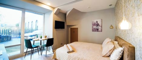 A modern bedroom with a large bed, a dining area with chairs, a table, and a view of the outside.