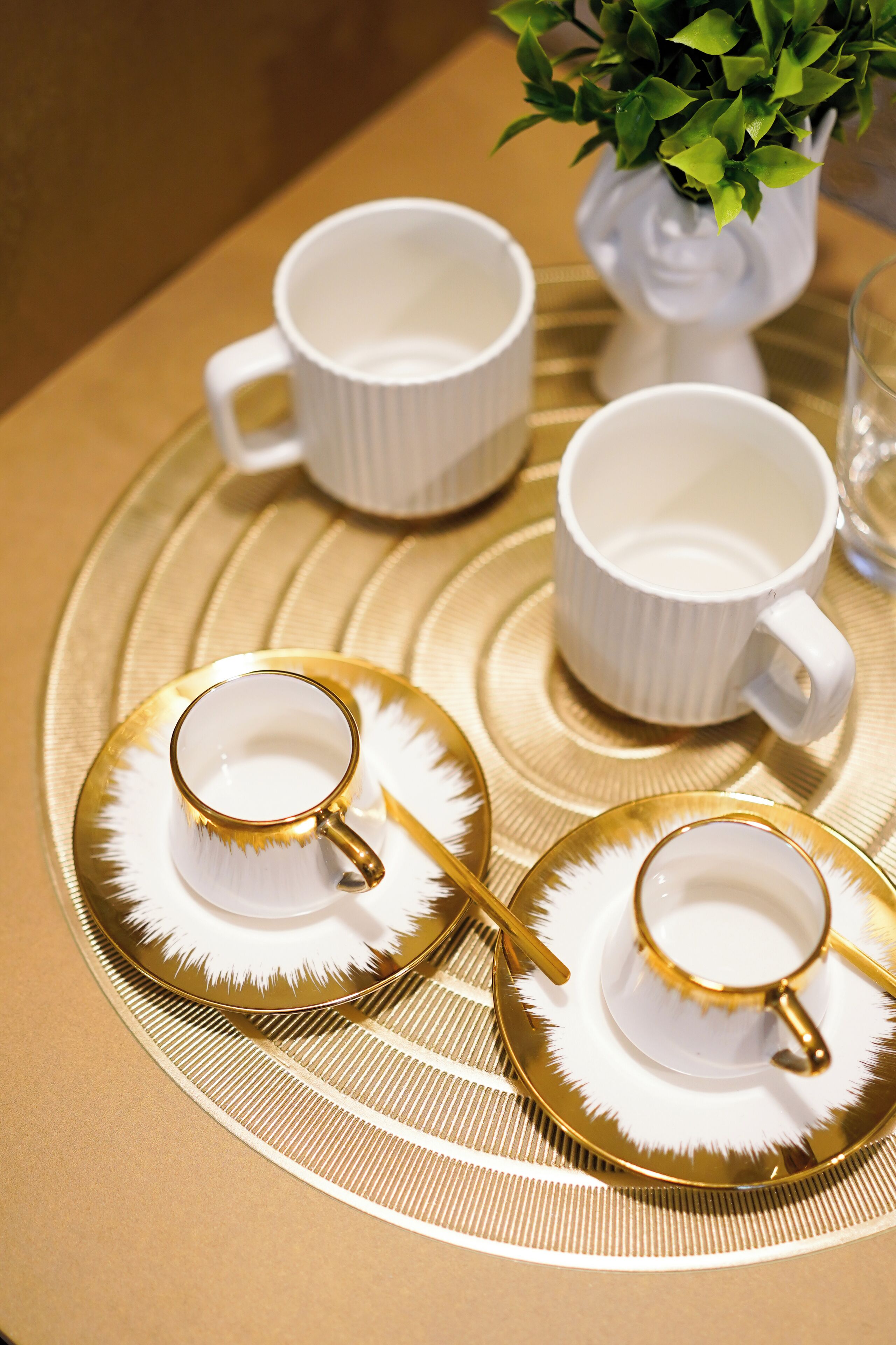 A table setting with white cups and gold-rimmed saucers, a white vase with green plants, and a gold tray.
