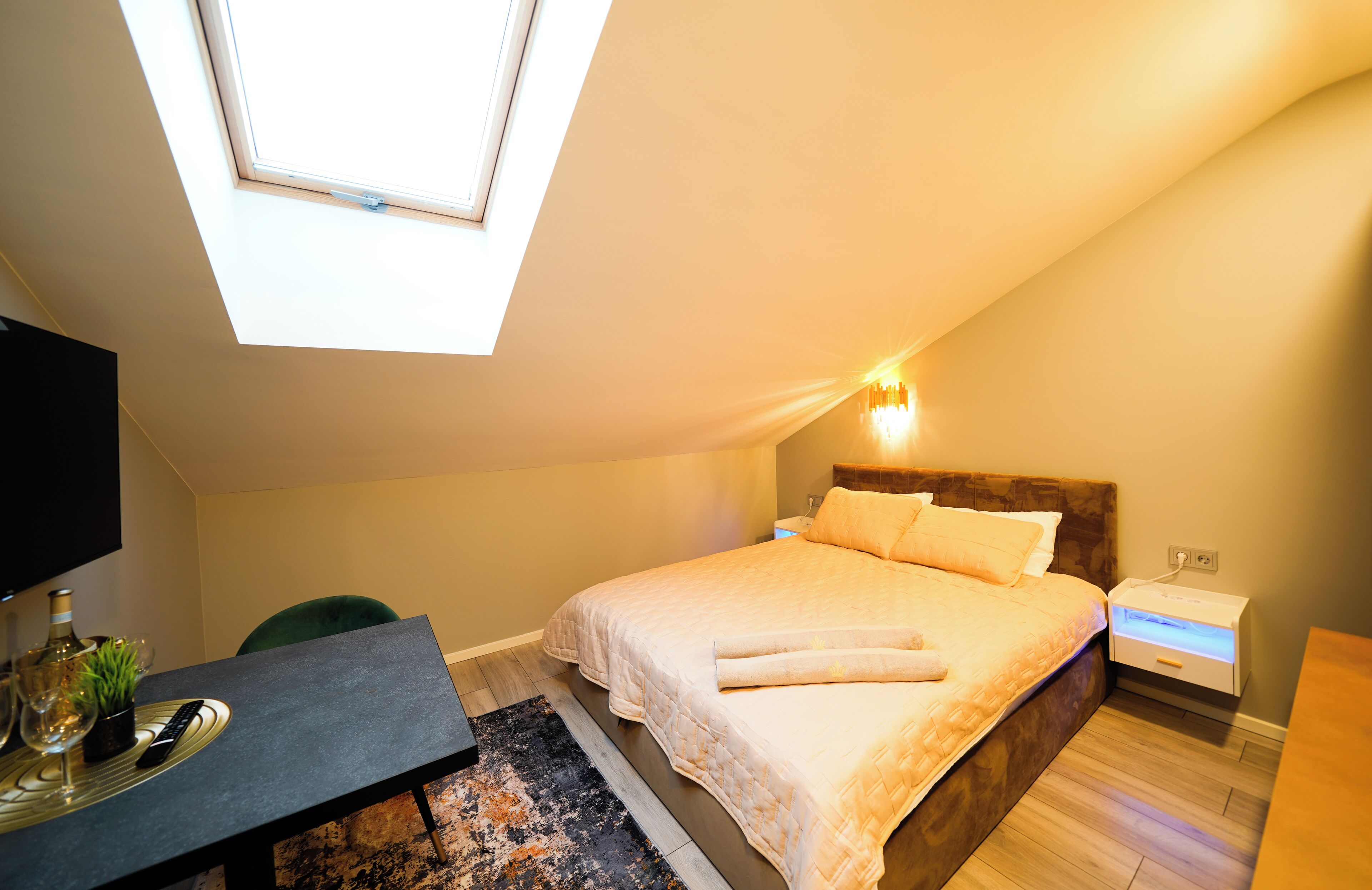 Un dormitorio con tragaluces, una cama con cabecera de madera, mesitas de noche, un escritorio con una silla y una pequeña mesa con una planta y un control remoto.