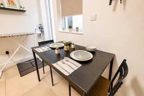 Ein Essbereich mit einem dunklen Holztisch, gedeckten Plätzen für zwei Personen, weißen Tellern, Besteck und einem kleinen Topfpflanzen.