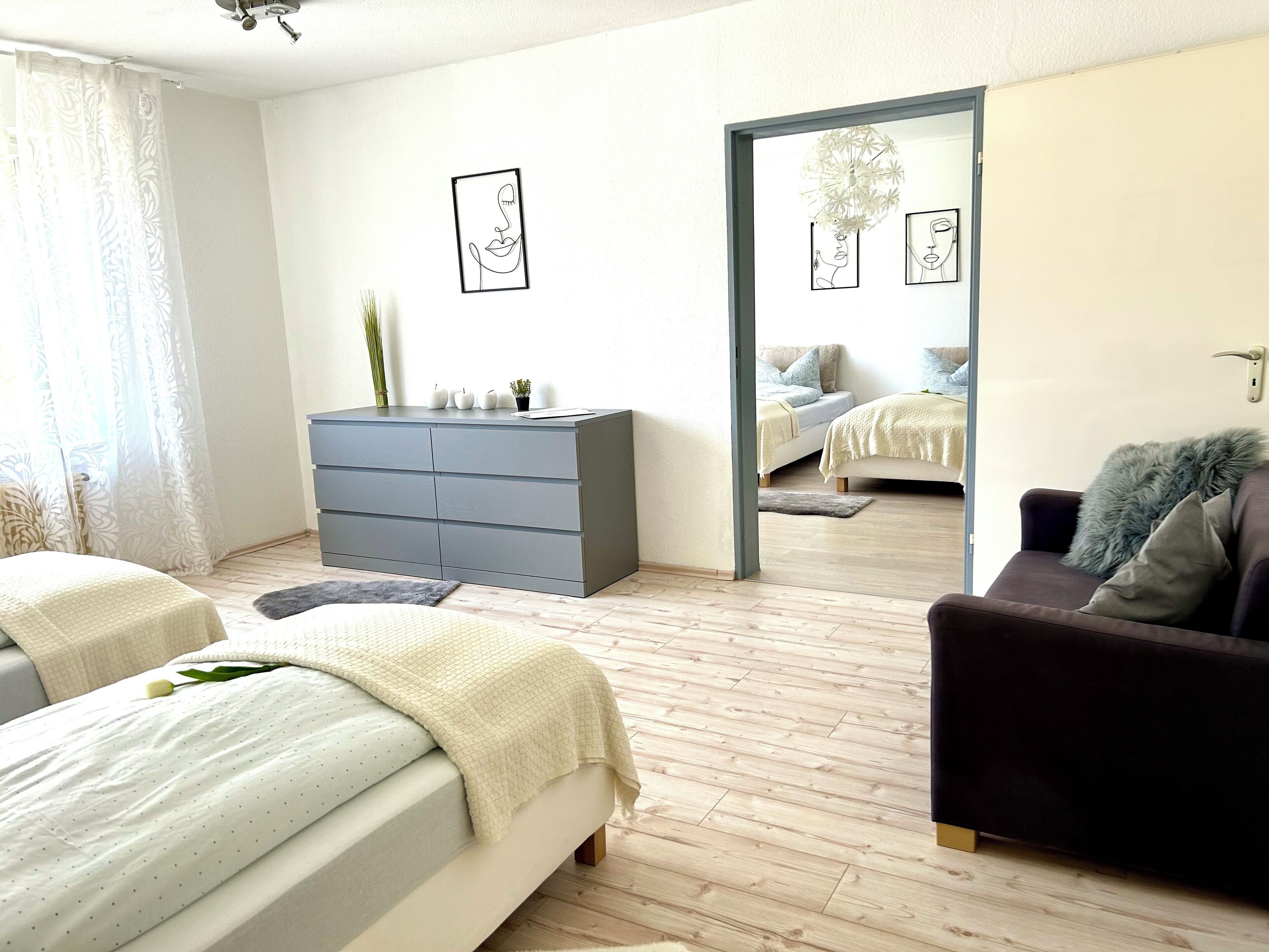 A modern bedroom with a large bed, a dresser, and a sofa. There are framed artworks on the wall and a view into another room through an open door.