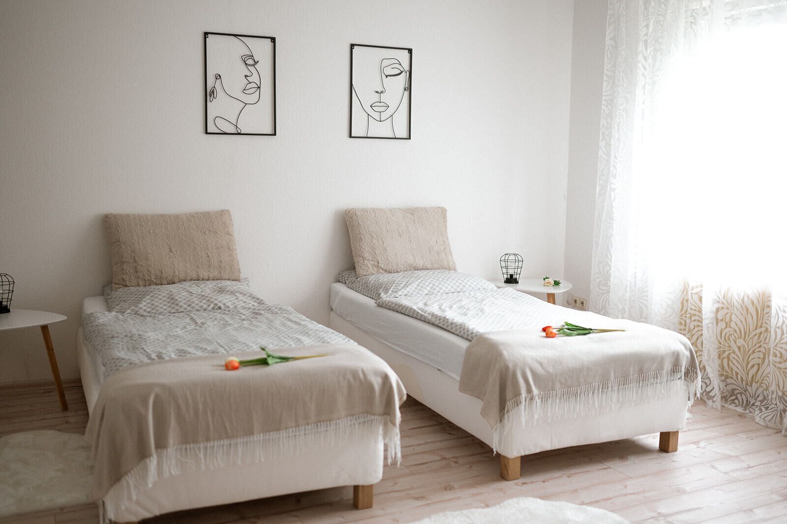 Een minimalistische slaapkamer met twee bedden, een klein tafeltje en twee ingelijste kunstwerken aan de muur.