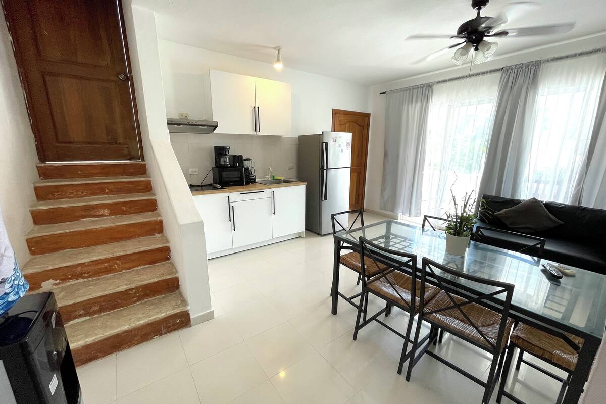 A modern kitchen with white cabinets, a built-in microwave, and a refrigerator. There's a glass dining table with chairs and a ceiling fan above.