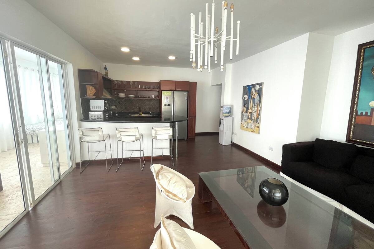 A modern living space with a kitchen, a dining area with bar stools, a glass coffee table, and a black sofa.