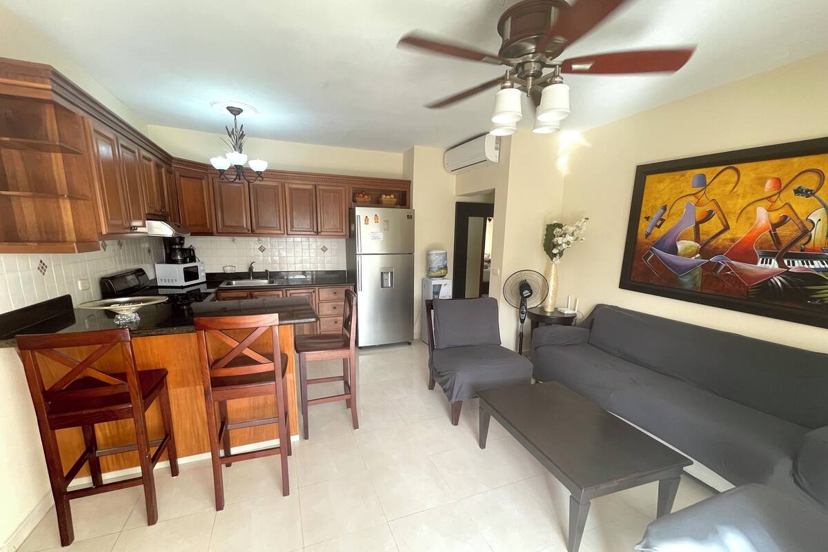 A modern kitchen with wooden cabinets, a dining area with a table and chairs, a gray sofa, and a ceiling fan.