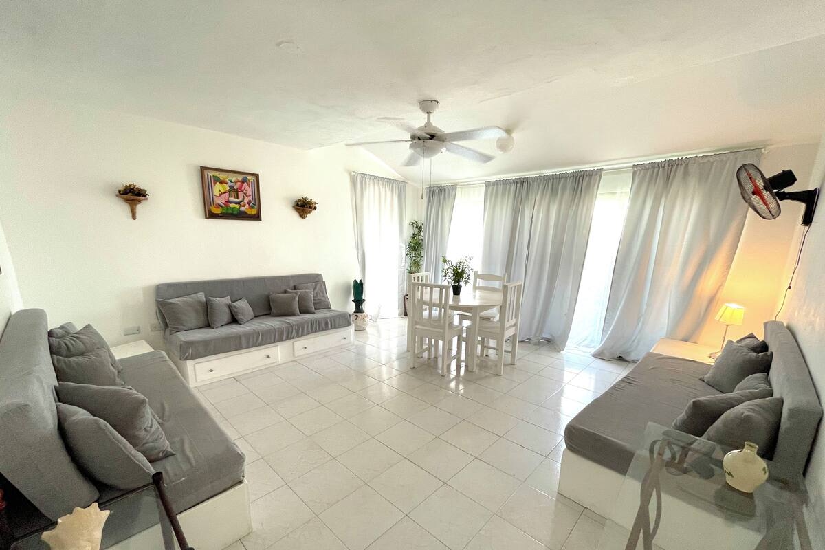 A spacious living area with a grey sofa, a dining table with chairs, a ceiling fan, and a large window with curtains.