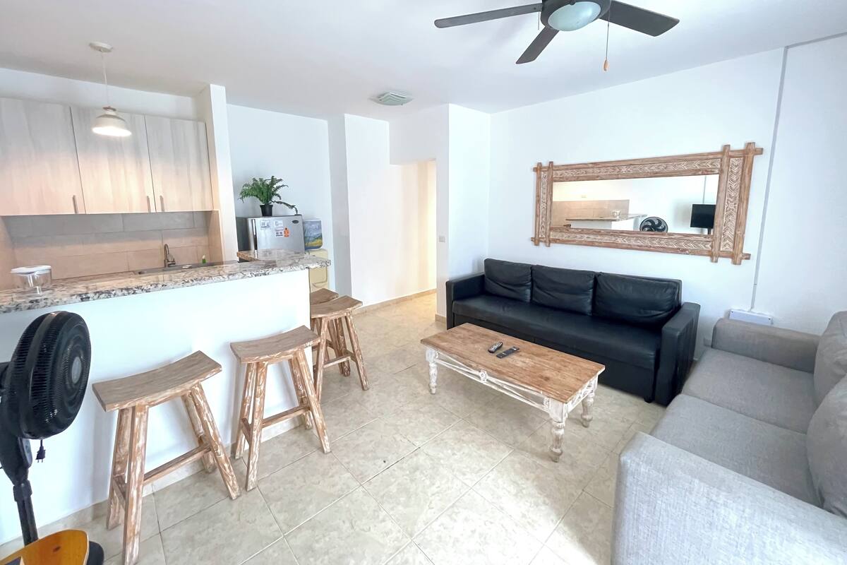 A modern living space with a kitchen, a black leather sofa, a wooden coffee table, and a ceiling fan.