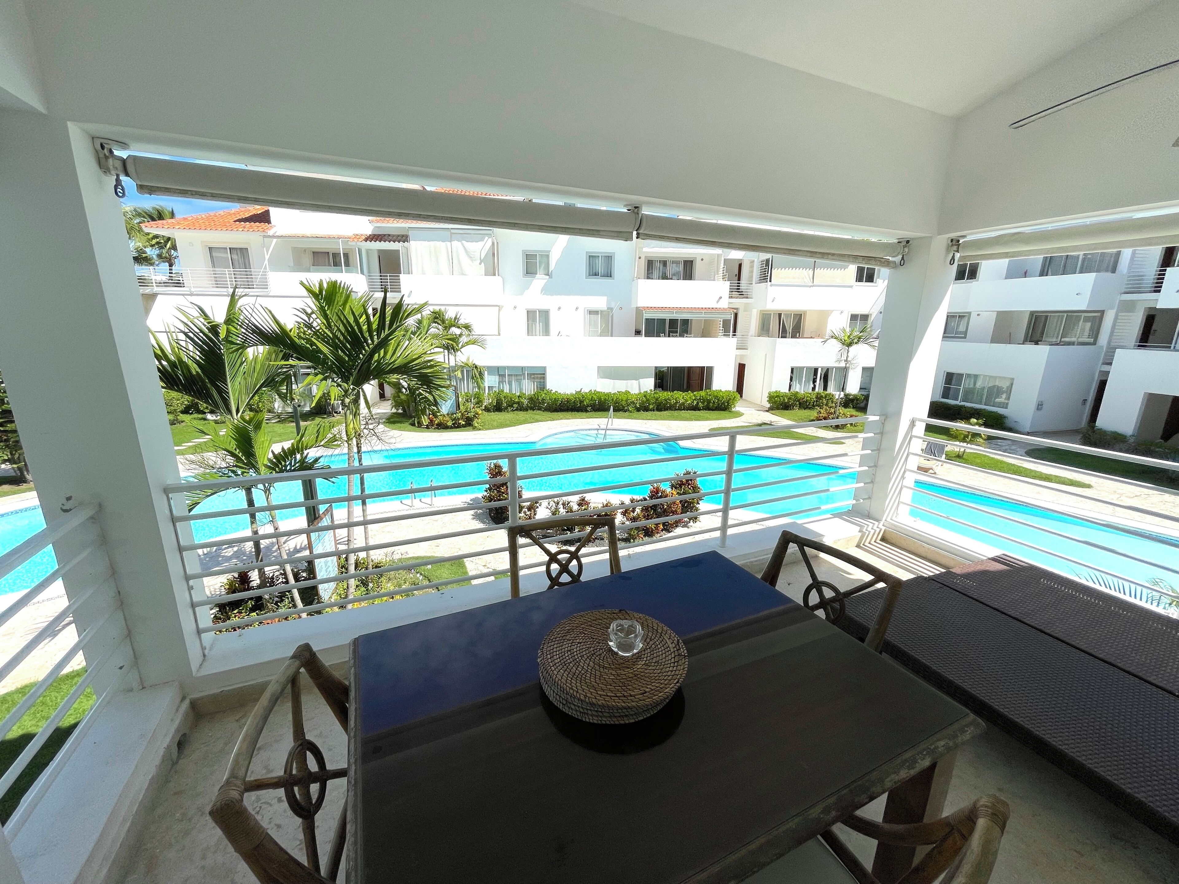 A balcony with a view of a swimming pool, white buildings, and greenery.