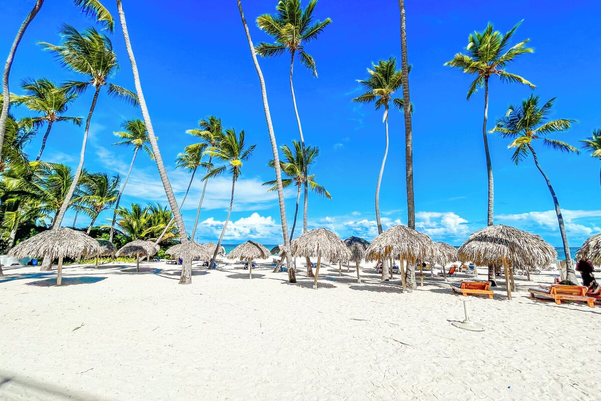 A beach with palm trees and thatched umbrellas.