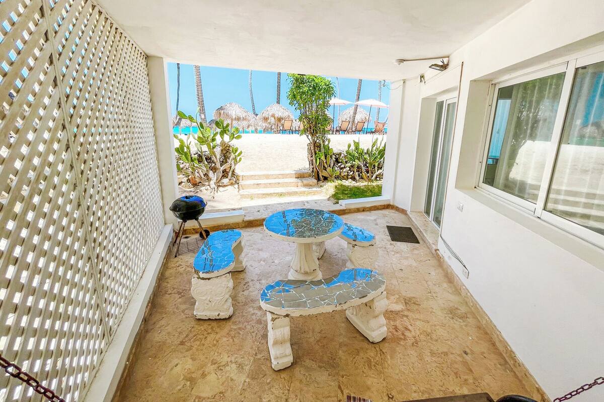 A balcony with a view of a beach, a grill, and a mosaic table.