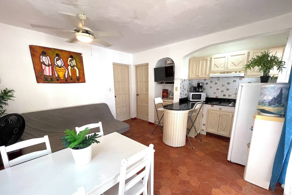 A compact kitchen with white cabinets, a dining table with chairs, a ceiling fan, and a painting on the wall.