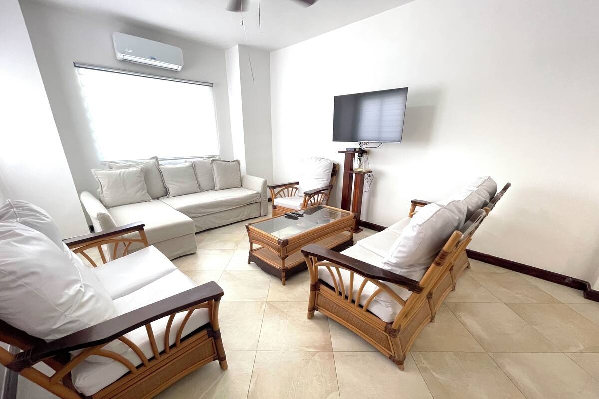 A living room with a sofa, two wooden chairs, a coffee table, and a flat-screen TV mounted on the wall.