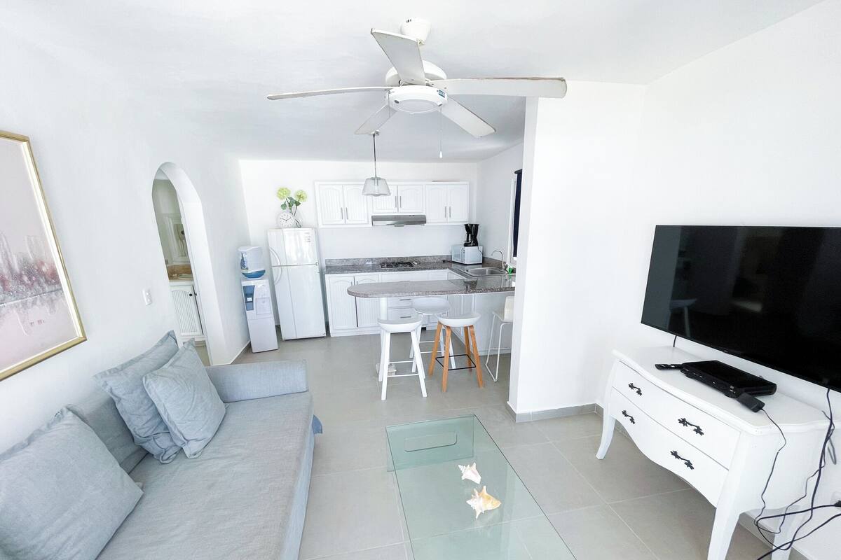 A modern living room with a sofa, a glass coffee table, a TV, and a kitchen in the background.