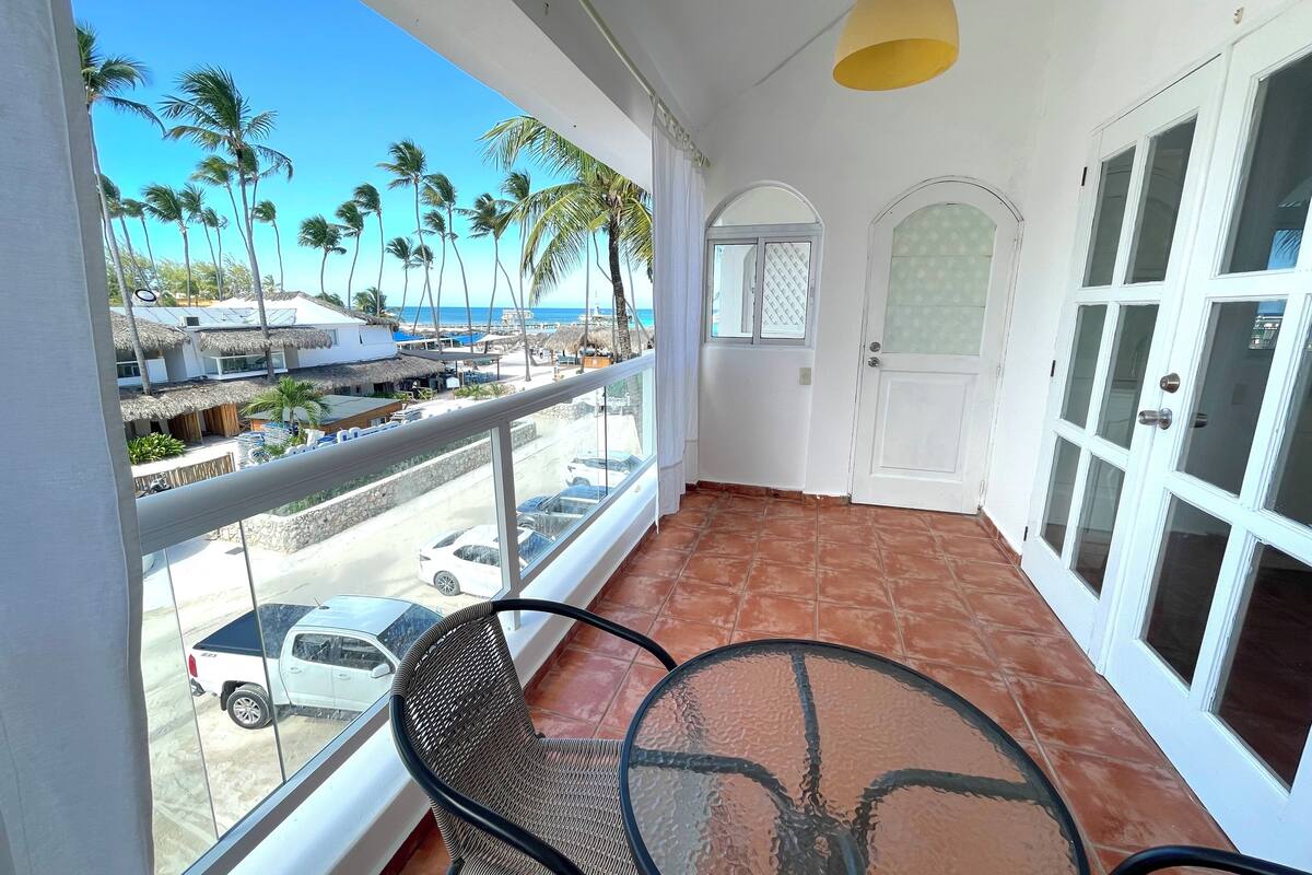 A balcony with a glass table and chairs, a white door, and a view of a beach with palm trees and parked cars.