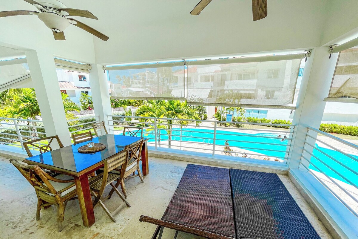 Un área junto a la piscina con mesa y sillas, un ventilador de techo y vistas a una piscina.