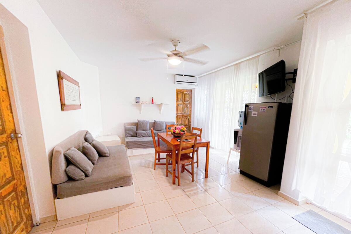 A living room with a sofa, a dining table with chairs, a ceiling fan, and a refrigerator.