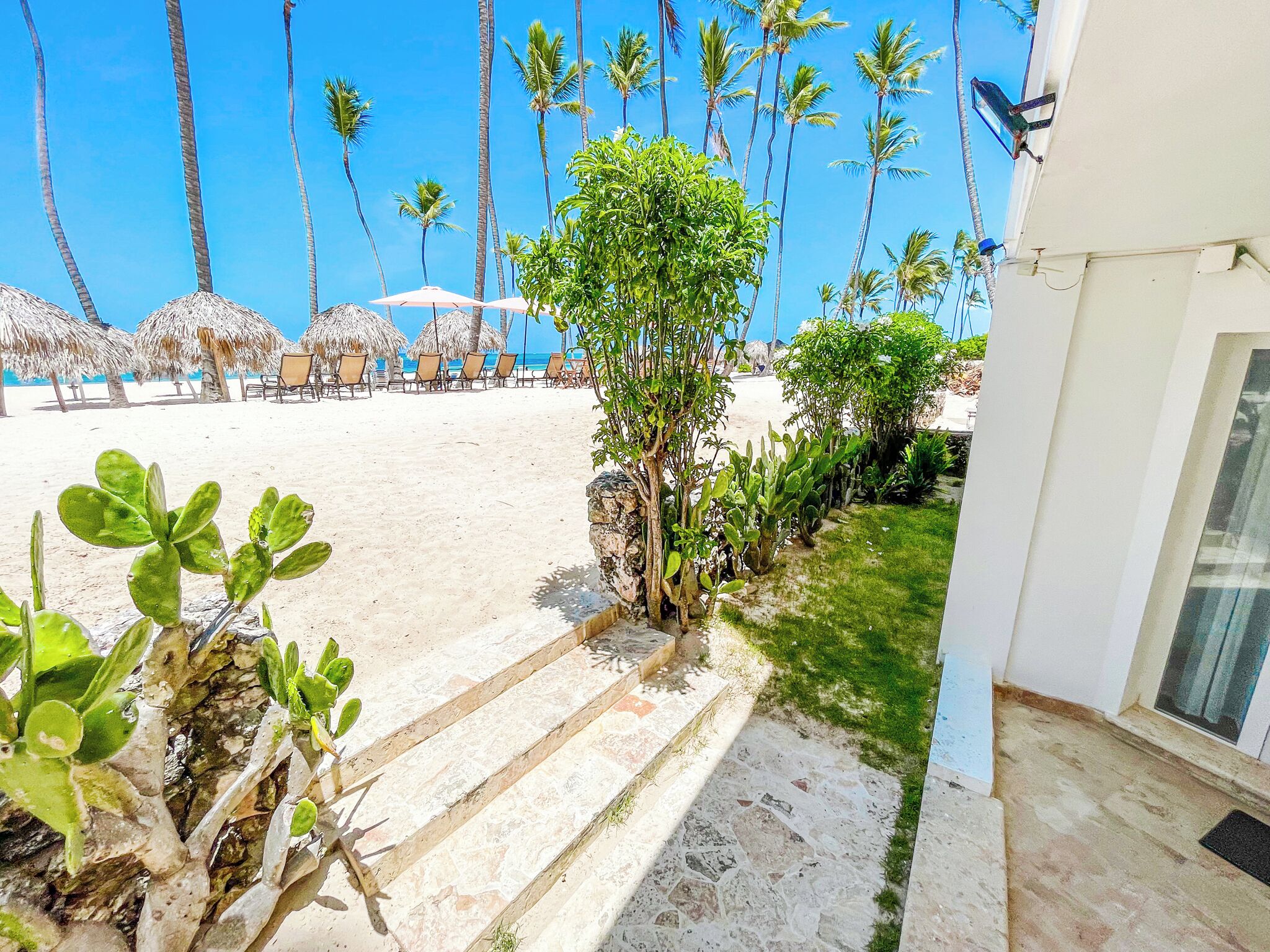 A beachside hotel with a stone staircase leading to a sandy area, palm trees, and beach chairs.