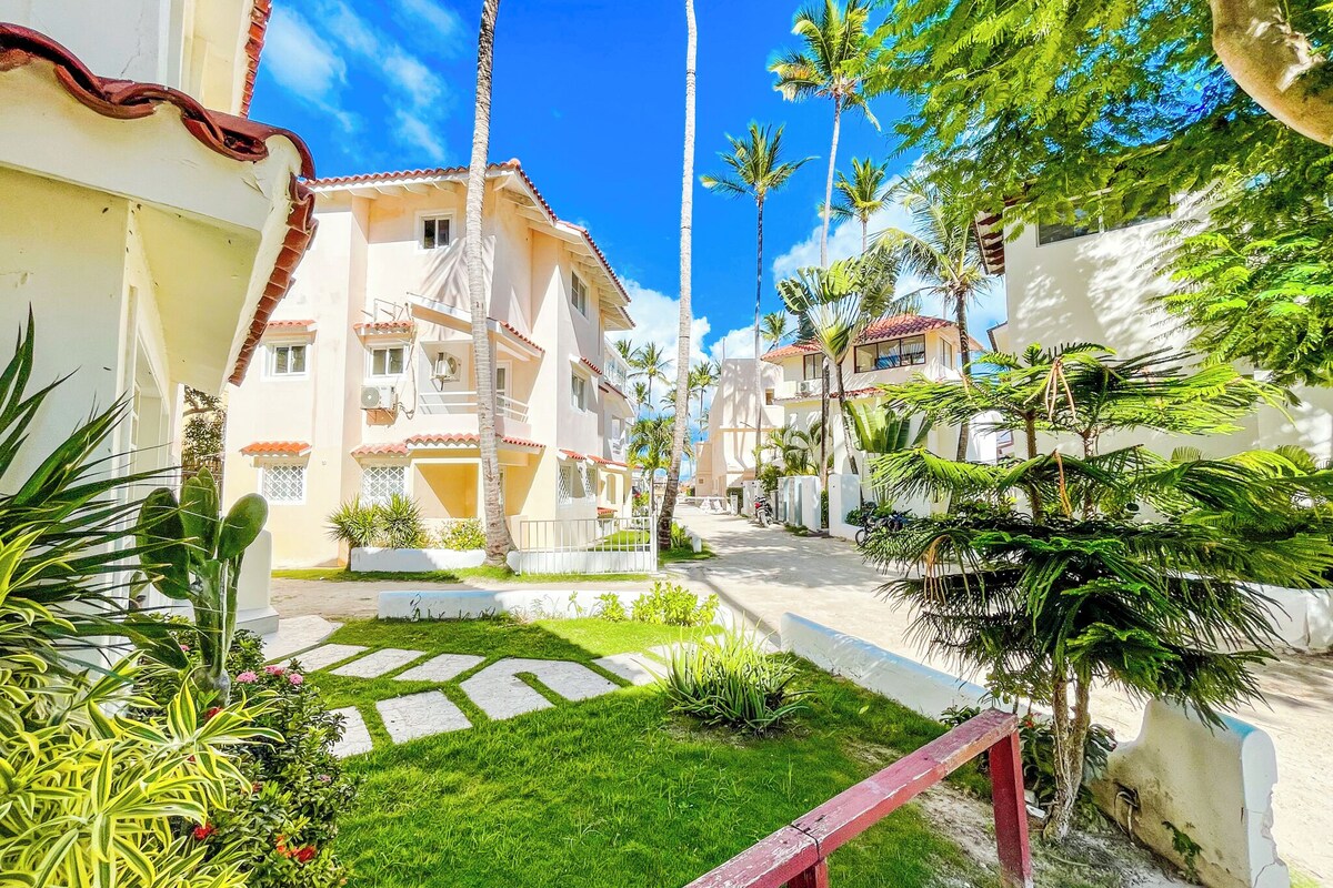 A residential area with a paved pathway, palm trees, and a building with a tiled roof.