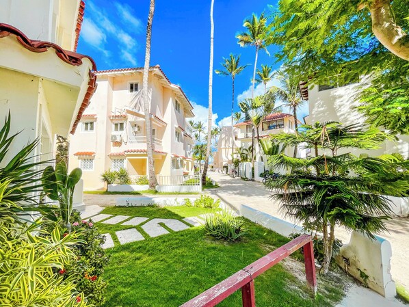 A residential area with a paved pathway, palm trees, and a building with a tiled roof. - MINI BEACH STUDIOS (Punta Cana)