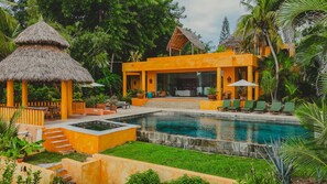 A resort with a swimming pool, a thatched-roof structure, and a modern yellow building with outdoor seating. - Casa Tara - Luxury Beach Escape in Punta de Mita (Punta de Mita)