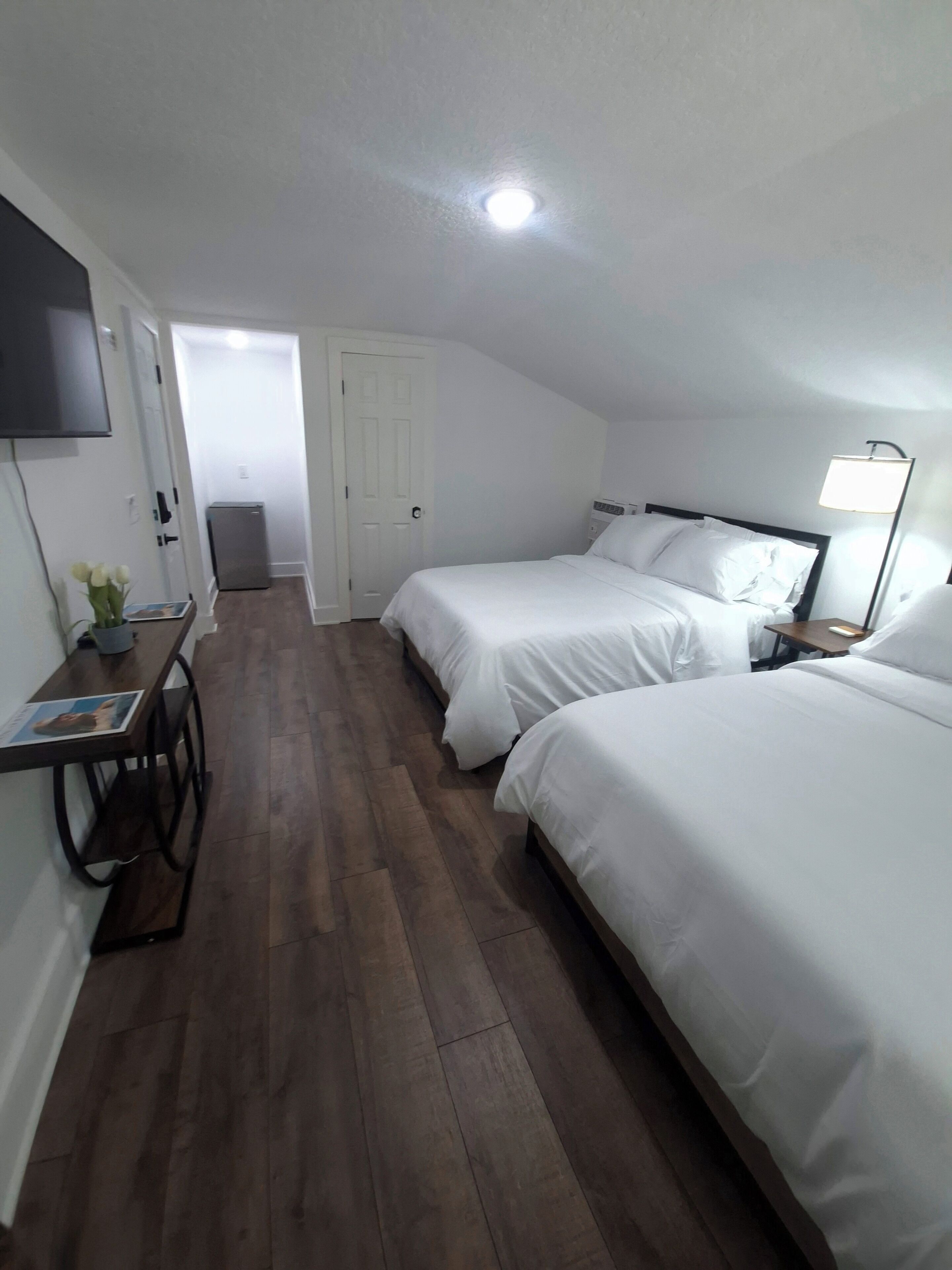 Habitación de hotel con dos camas, un escritorio con silla, televisor montado en la pared y puerta que da a otra habitación.