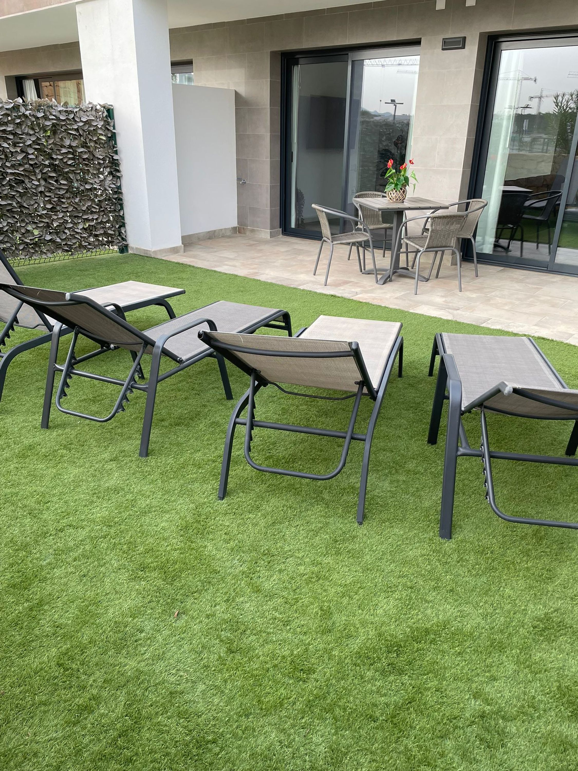 An outdoor seating area with metal chairs and tables, a glass door leading to a patio, and a wall covered with foliage.