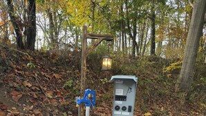 A utility box with a control panel, a blue clamp, and a white object on a gravel surface with fallen leaves. - 50/30 amp all weather campground site #2 full hook up (Masury)