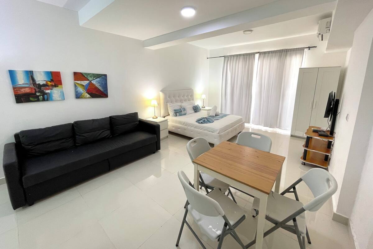 A modern, well-lit room with a black sofa, a wooden dining table with chairs, a bed with a white headboard, and a wardrobe.