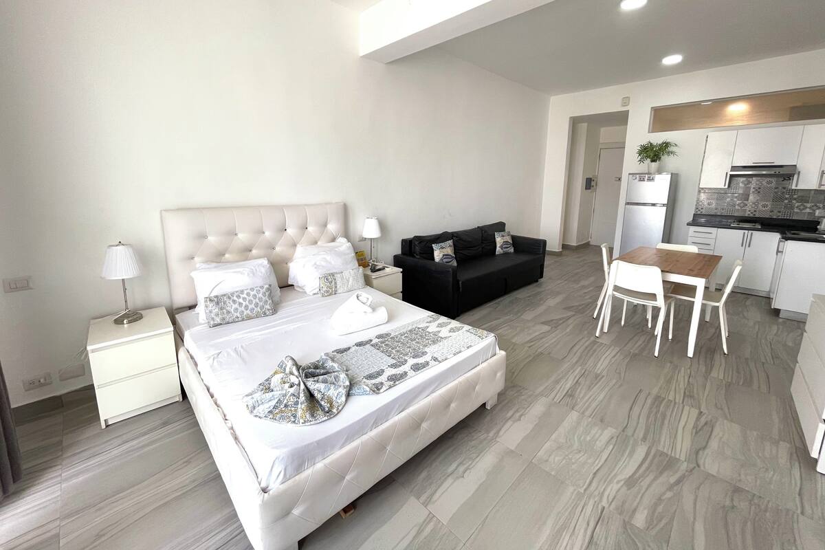 A modern hotel room with a large bed, a sofa, a dining table, and a kitchenette.