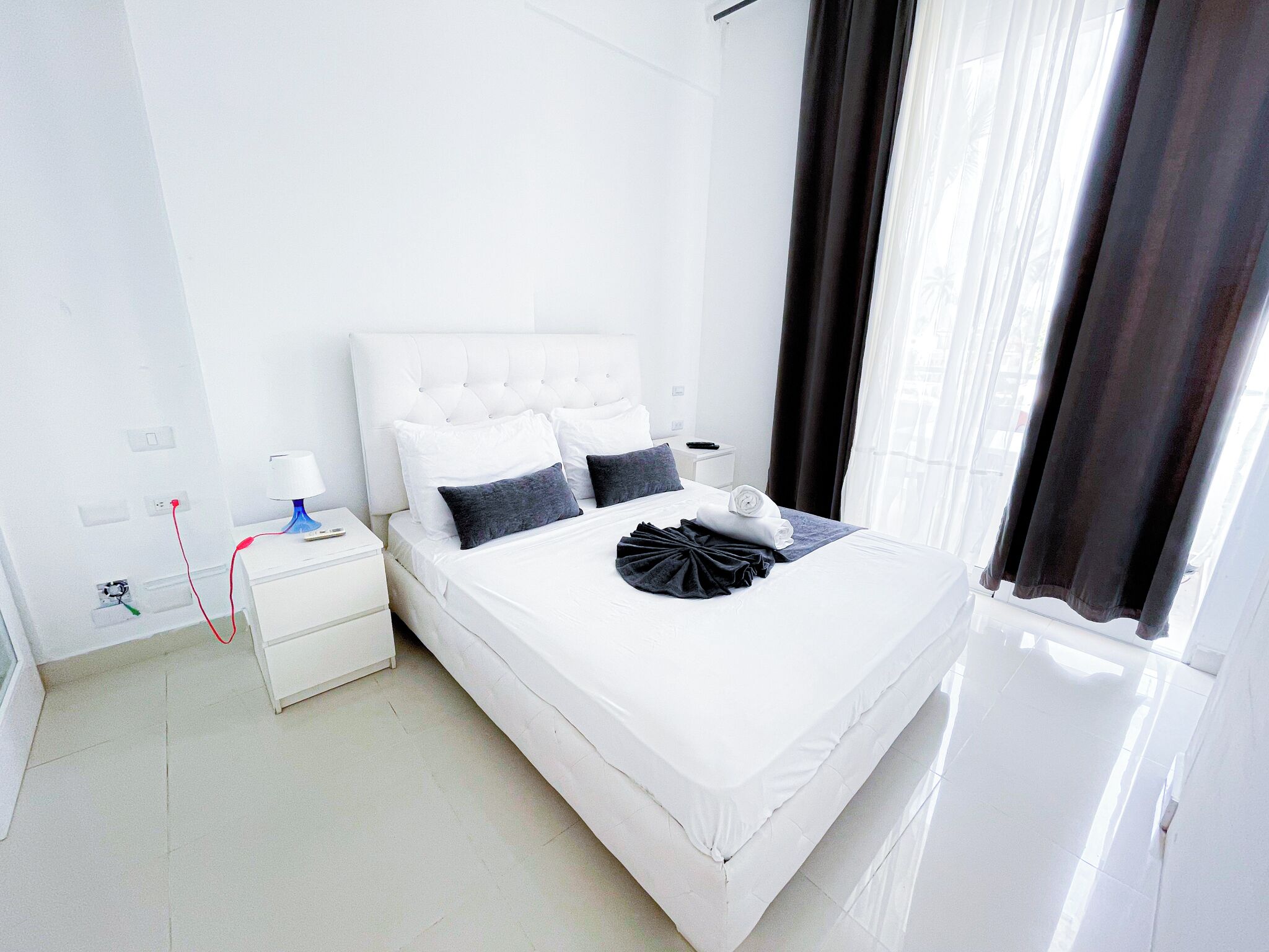 Une chambre d’hôtel avec un lit blanc, une table de chevet avec une lampe et une fenêtre avec des rideaux.