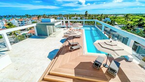 A rooftop pool area with lounge chairs and umbrellas, surrounded by a cityscape and greenery. - MINI STUDIOS BEACH (Punta Cana)