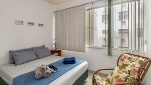 Quarto com cama, uma cadeira, janela com persianas e mesa de cabeceira com abajur.