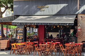 A restaurant with outdoor seating, a sign reading "Restaurant Novo," and a banner advertising a happy hour with a beer offer. - Plaza Copacabana - Conforto e Praia. (Rio de Janeiro)