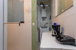 Una kitchenette pequeña con cafetera, hervidor de agua y plancha portátil sobre la encimera.