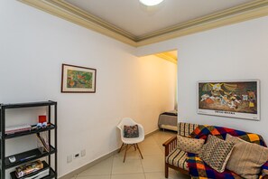 A living room with a sofa, a white chair, a bookshelf, and two framed artworks. - Plaza Copacabana - Conforto e Praia. (Rio de Janeiro)