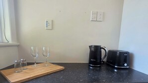 A kitchen with a wooden cutting board, two wine glasses, salt and pepper shakers, a black electric kettle, and a toaster on the counter.