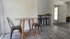 A dining area with a round wooden table and four chairs, a kitchen area with a countertop and stools, and a hallway leading to another room.
