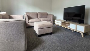 A modern living room with a beige sofa, a matching ottoman, a television on a wooden stand, and an air conditioning unit on the wall.